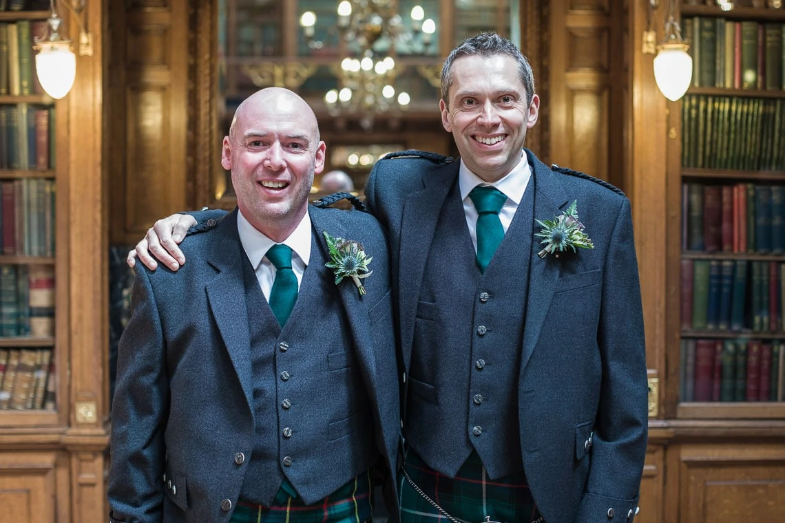 Two smiling men in traditional scottish attire with kilts and tweed jackets, standing arm-in-arm in a library with ornate woodwork and a chandelier.