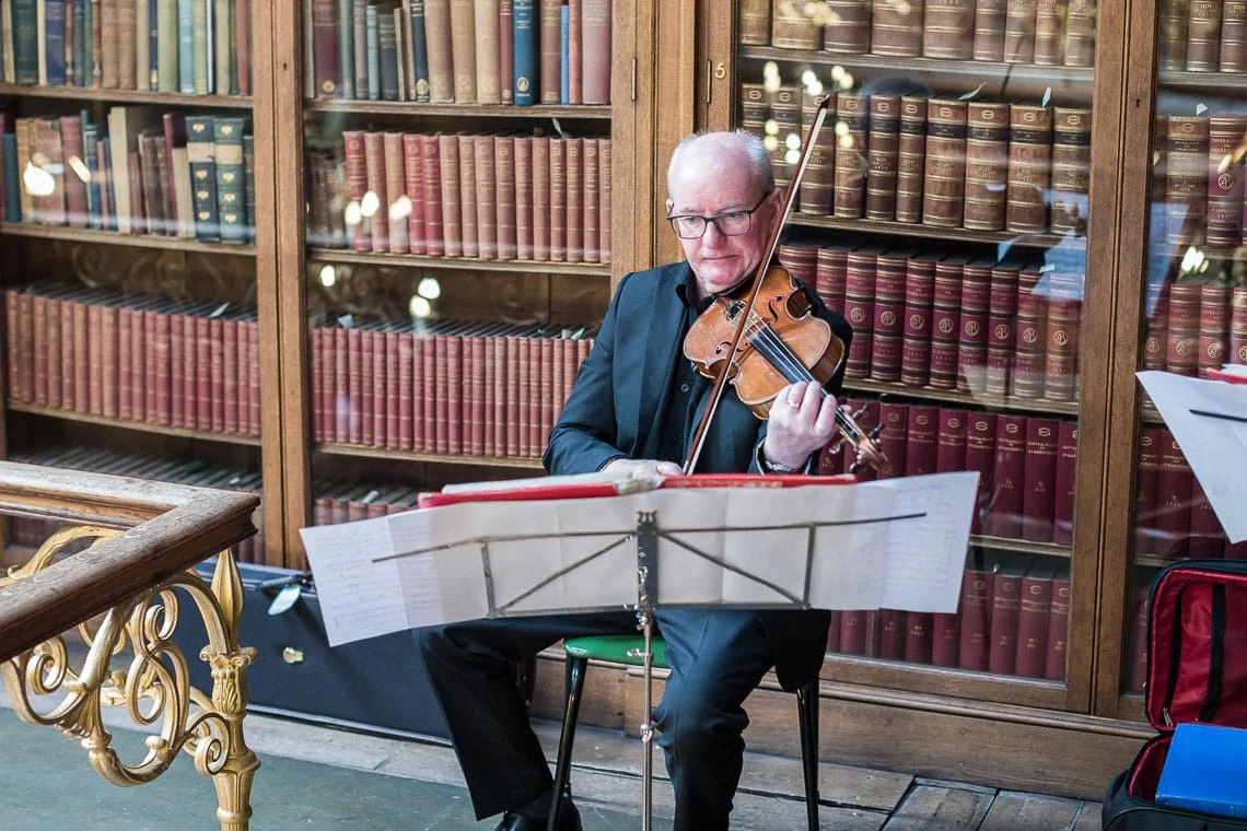 An elderly man in a black suit playing a violin in a library filled with books, focusing intently on sheet music on a stand.