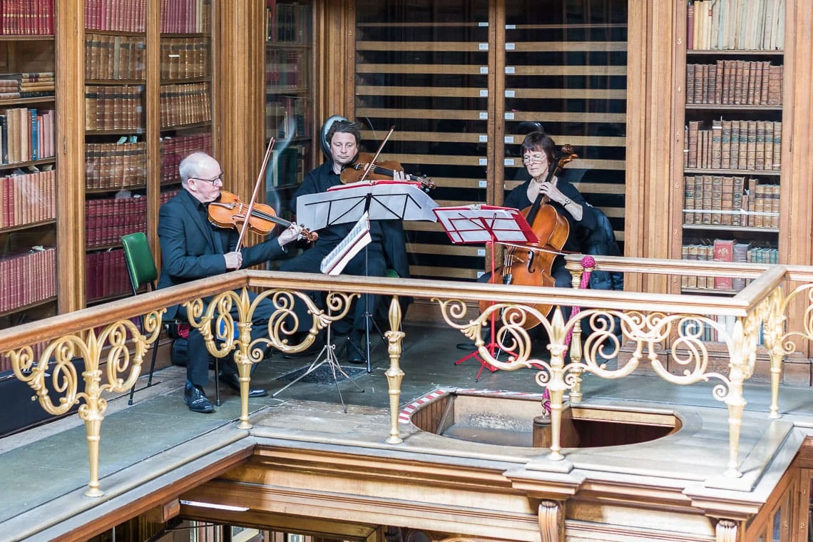 Three people performing a classical music piece, two on violins and one on a cello, surrounded by books in an ornate library setting.