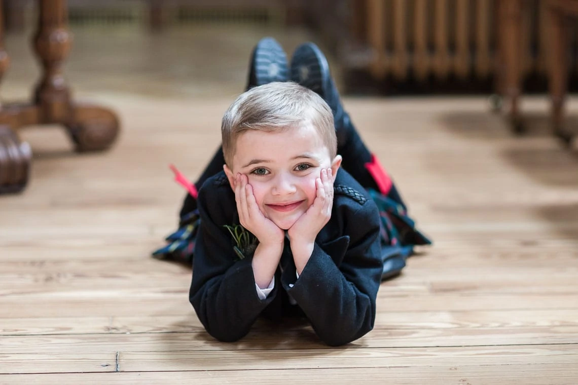 A young boy lying on a wooden floor, smiling at the camera with his hands on his cheeks.