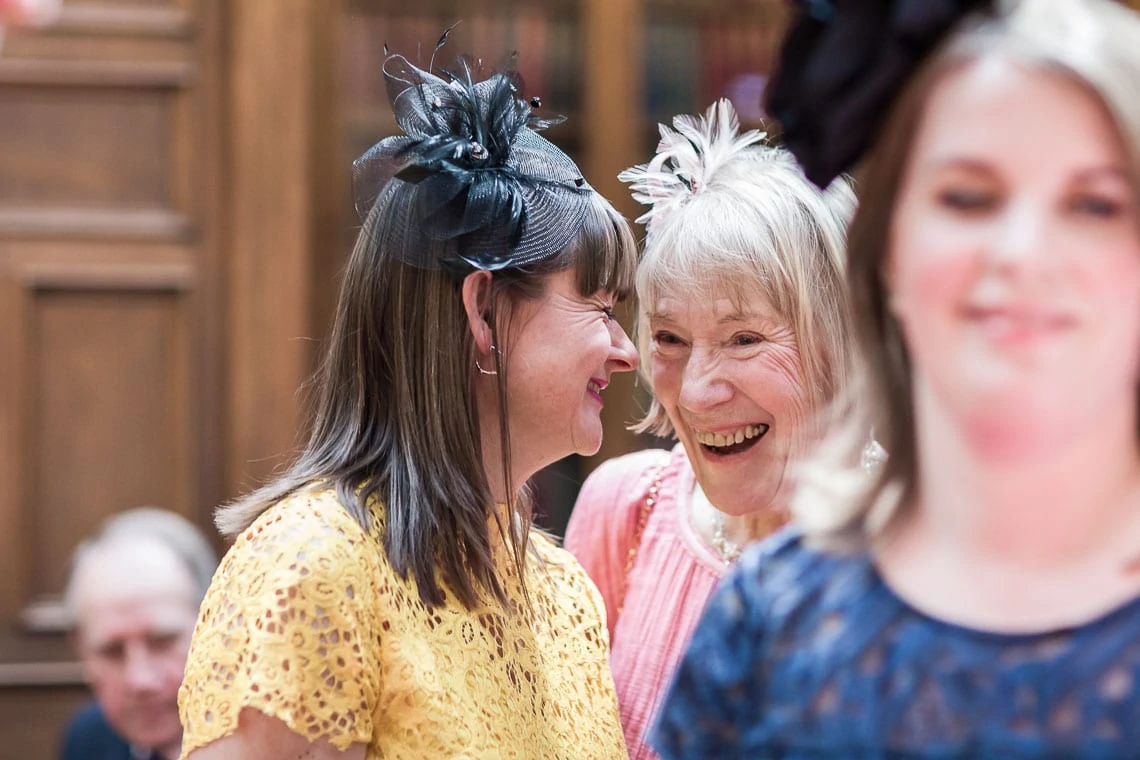 Two women smiling joyfully at one another, both wearing decorative fascinators, in a crowded indoor setting.