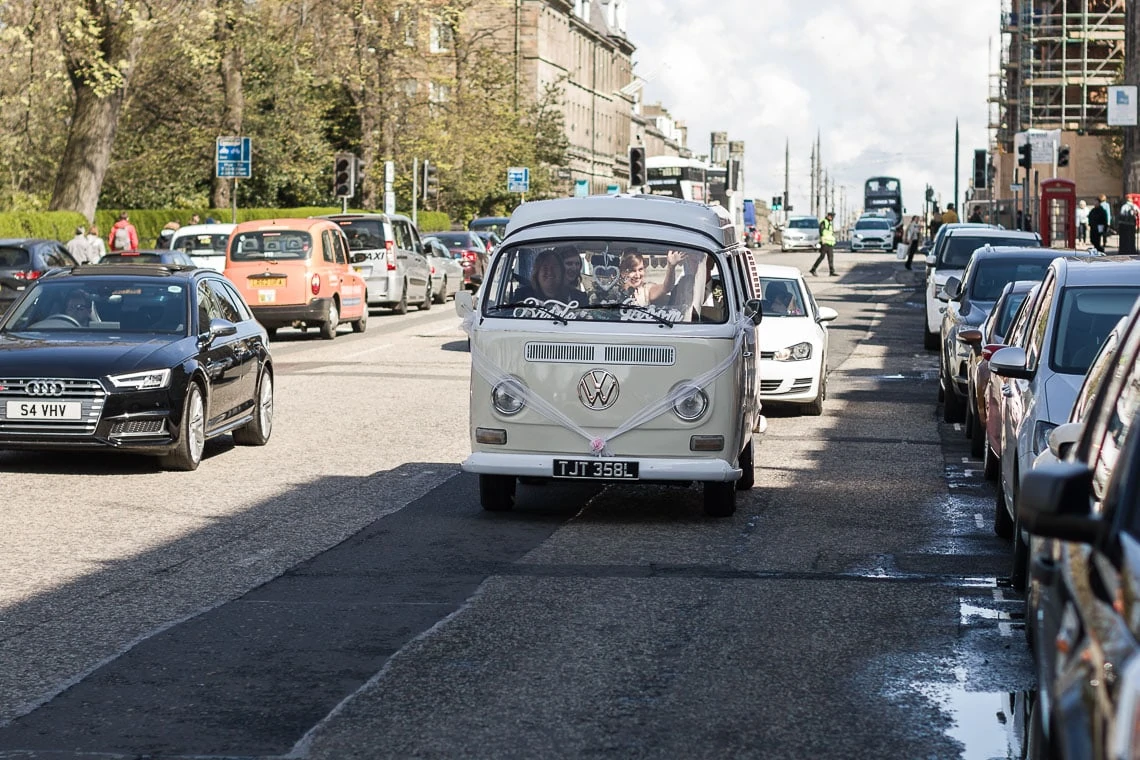 A vintage white volkswagen bus driving down a busy street lined with parked cars under a clear sky.