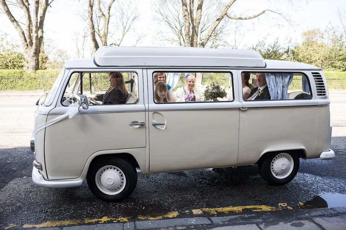 A vintage white volkswagen bus filled with people dressed in formal attire, parked on a city street on a sunny day.