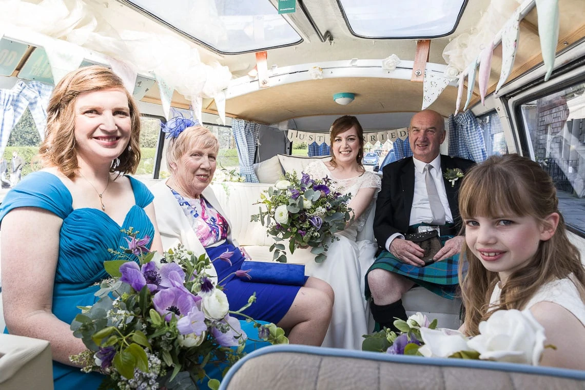 A wedding group sits inside a vintage van. one woman in blue holds a bouquet, accompanied by two women and a man in formal attire, and a young girl smiling at the camera.