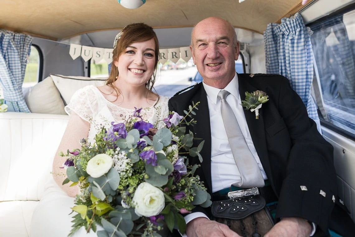 Bride holding a bouquet and sitting beside her father in a vintage car, both smiling, with a "just married" sign in the background.