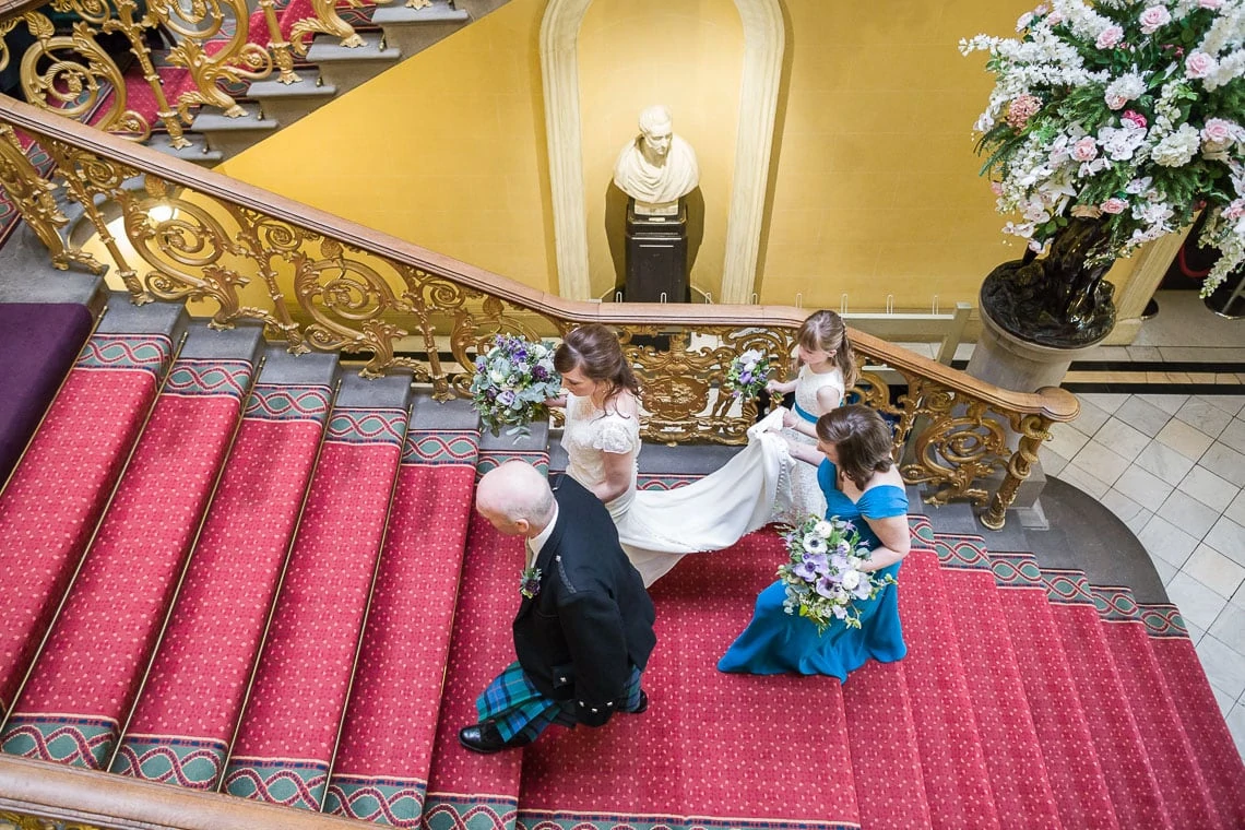 A bride in a white dress walks down a staircase assisted by a man in a kilt, followed by two bridesmaids in blue, inside an ornate building with floral decorations.