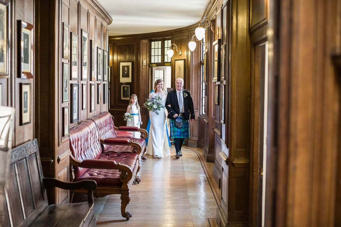 A bride and her father, the latter in a kilt, walk down a wood-paneled hallway with benches and framed pictures on the walls.