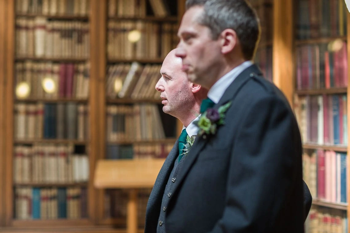 Two men in formal attire with boutonnieres stand side by side in a library with wooden shelves full of books.