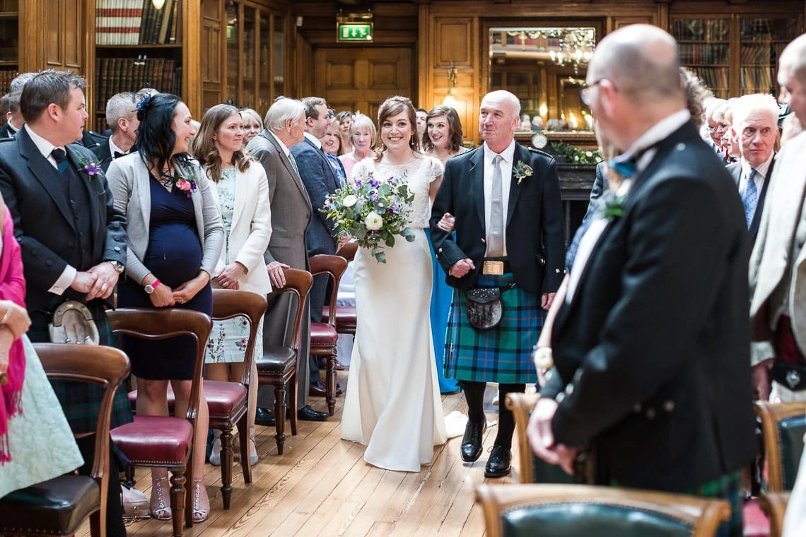 A bride walking down the aisle, smiling, accompanied by an older man in a kilt, with guests watching in a wood-paneled room.