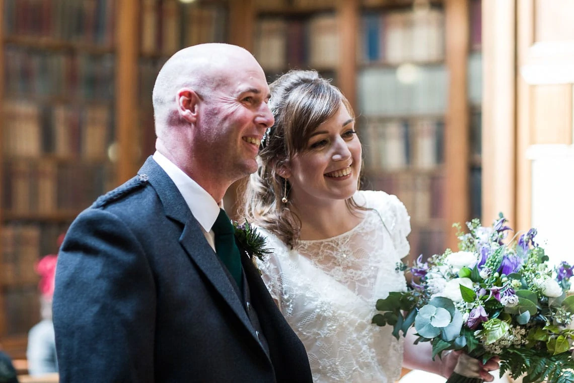 A smiling bride and groom holding a bouquet in a library with sunlight streaming through the windows.