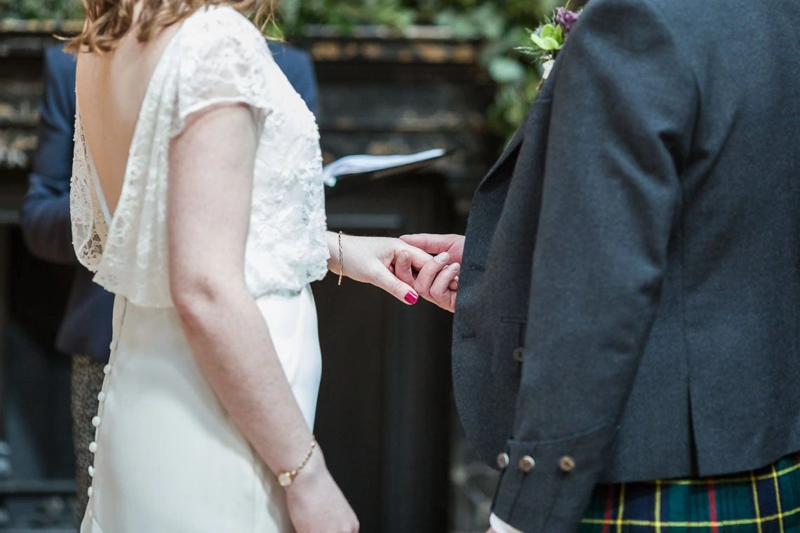 A bride in a white lace dress holds hands with a groom in a tartan kilt, symbolizing intimacy and connection at a wedding.