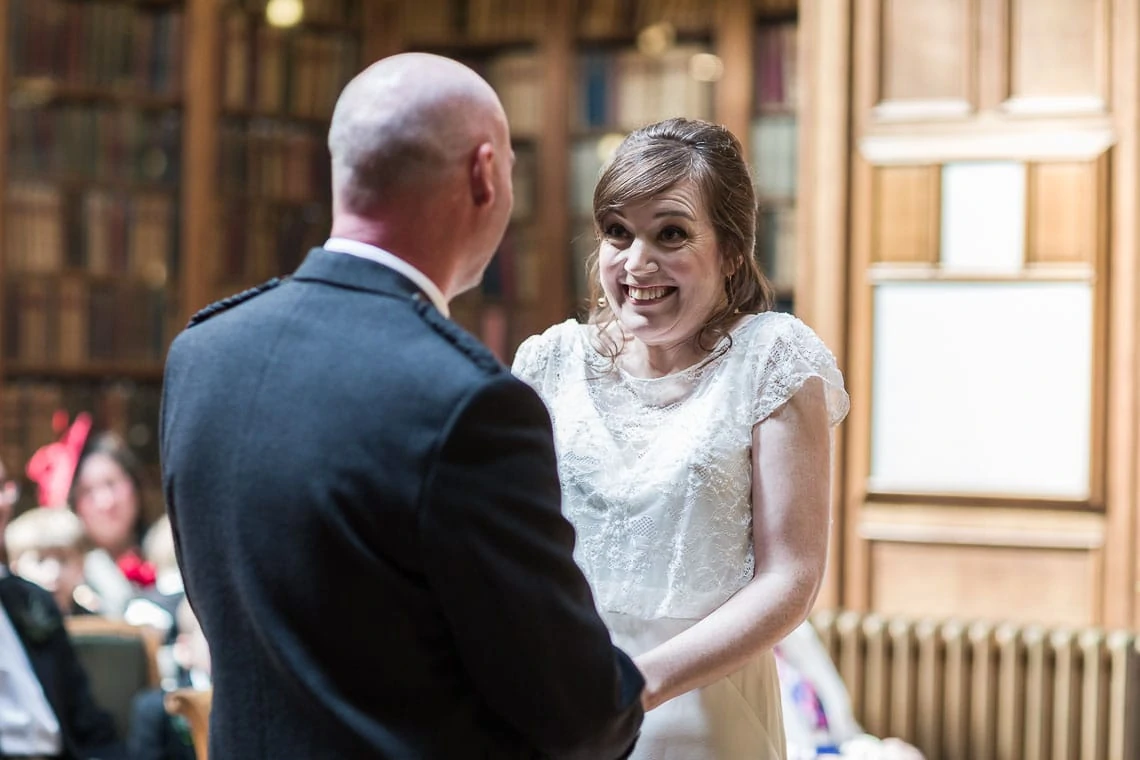 A bride in a white dress smiling joyfully at a groom in a formal suit during their wedding ceremony in a library with wooden shelves and guests in the background.
