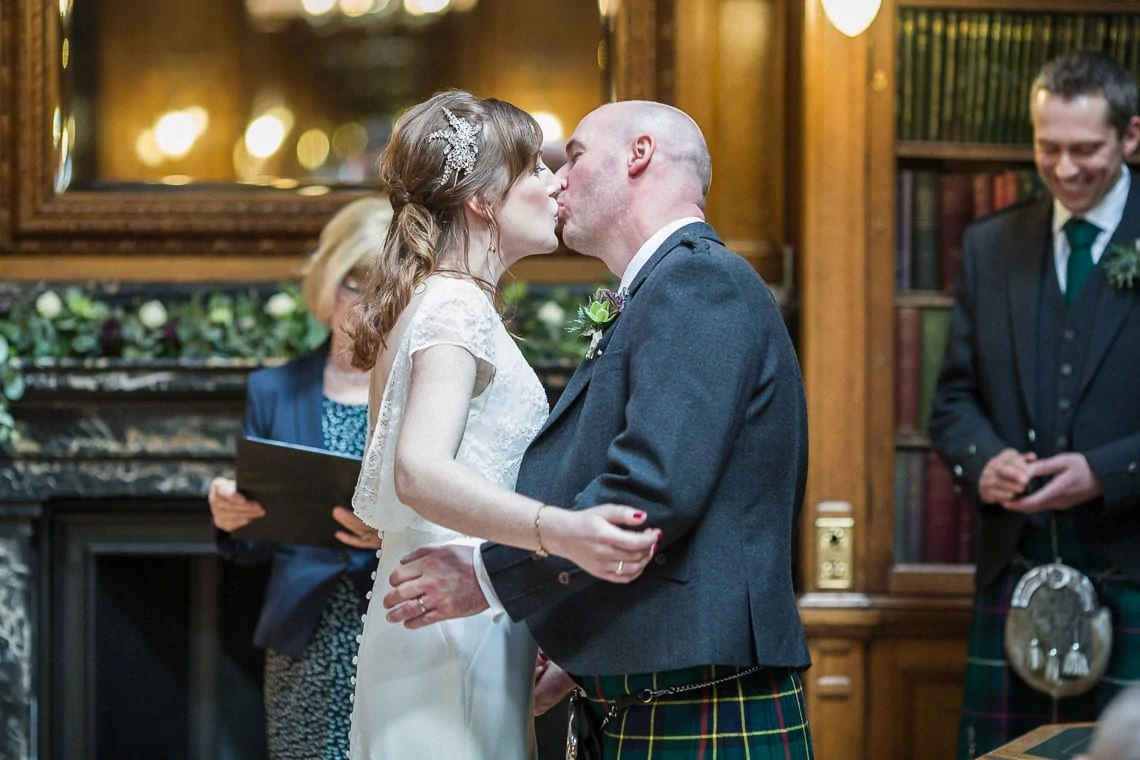A bride and groom kiss following their wedding ceremony, the groom wearing a kilt; an officiant and a smiling man are seen in the background.