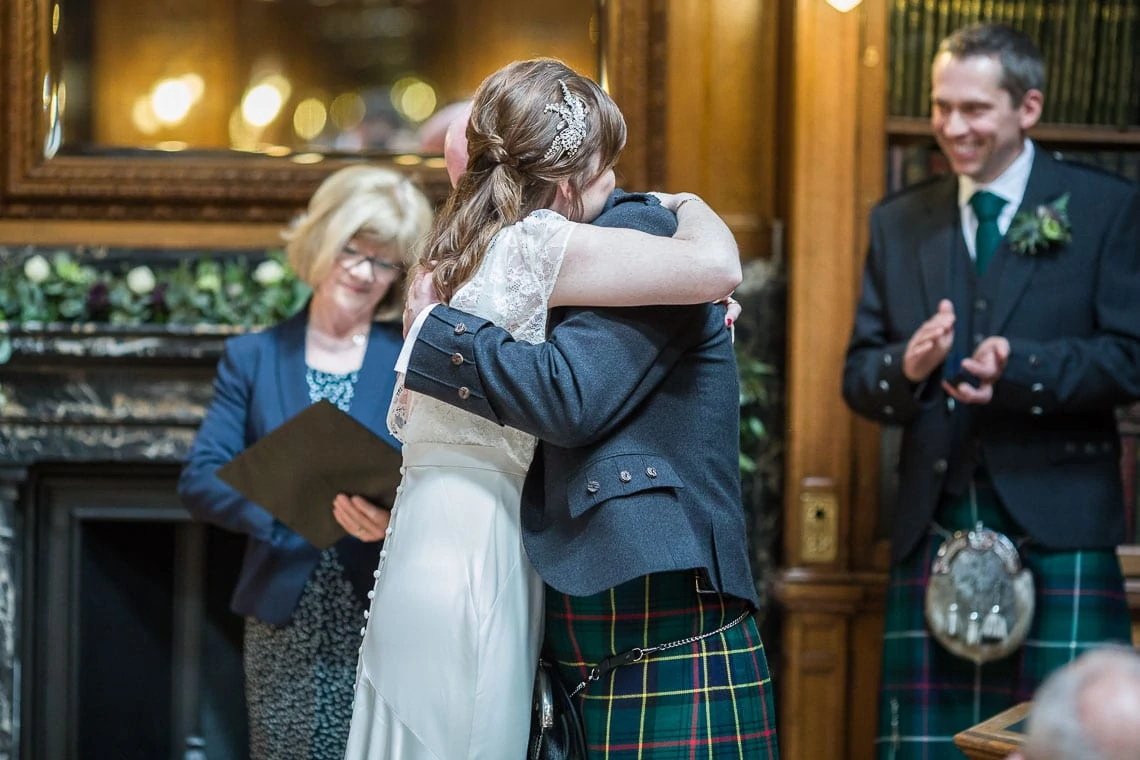 Bride and groom embracing at their wedding ceremony, with an officiant and a smiling man in a kilt observing.
