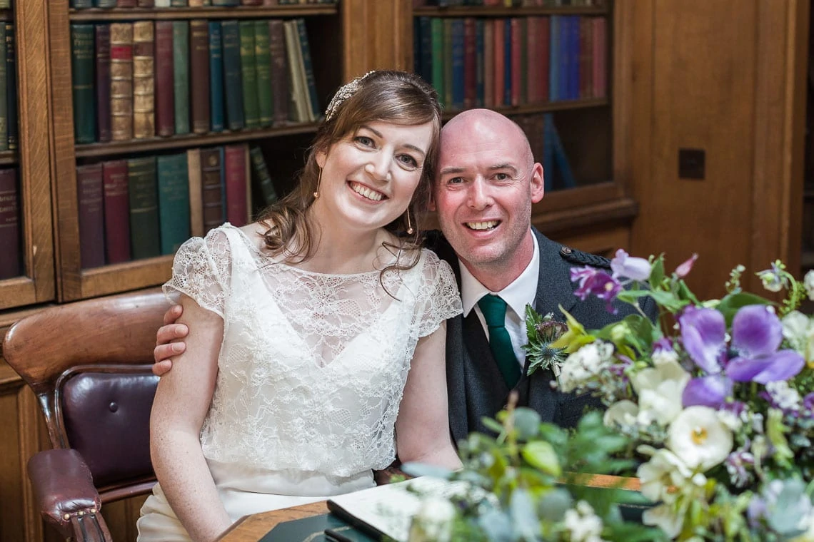A newlywed couple smiling joyfully, seated with a bouquet of purple flowers beside them, in a library setting.