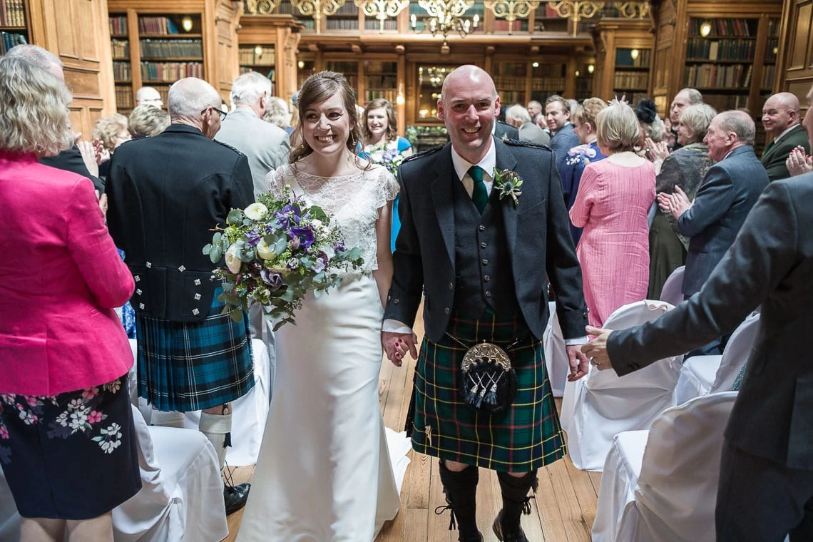 Bride and groom in scottish attire smile as they walk through a clapping crowd in an ornate library setting.