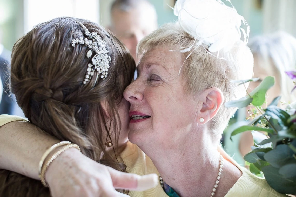 A young bride in a headpiece whispers to an elderly woman in a yellow outfit and white fascinator at a wedding.