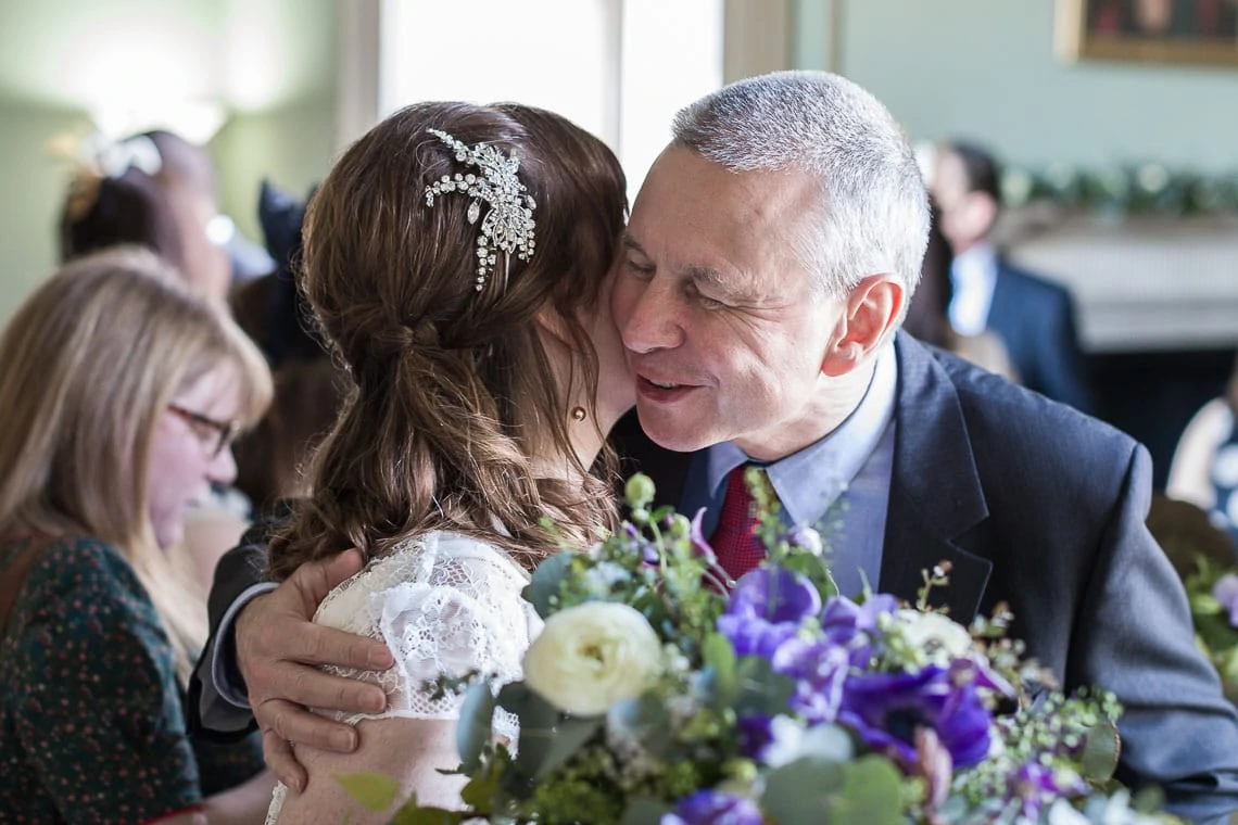 A bride and an older man embrace joyfully at a wedding reception, surrounded by guests and a table with purple floral arrangements.