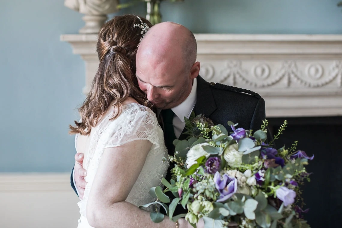 A bride and groom embracing affectionately, with the groom kissing the bride's head, surrounded by a decorative interior setting.