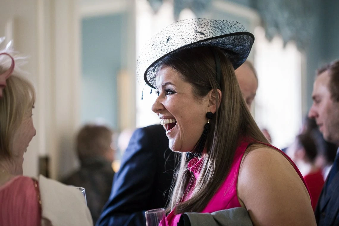 A woman in a pink dress and a stylish black and white hat laughs joyfully at a social event, holding a glass, surrounded by other guests.