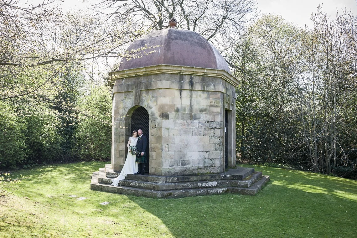 A bride and groom stand in the doorway of a small, domed stone pavilion surrounded by lush greenery on a sunny day.