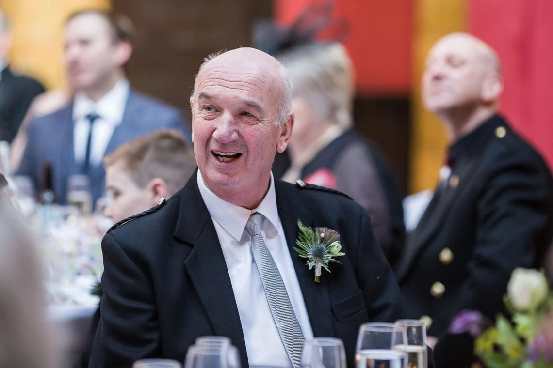 An older man in a formal suit with a flower boutonniere laughing at a table during a festive event.