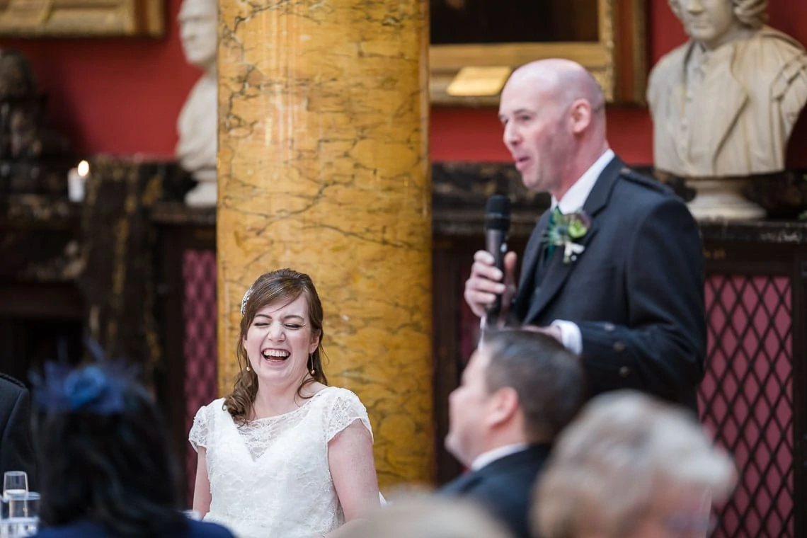 A bride laughs heartily as the groom speaks into a microphone during a wedding speech in an ornate room with marble columns and bust sculptures.