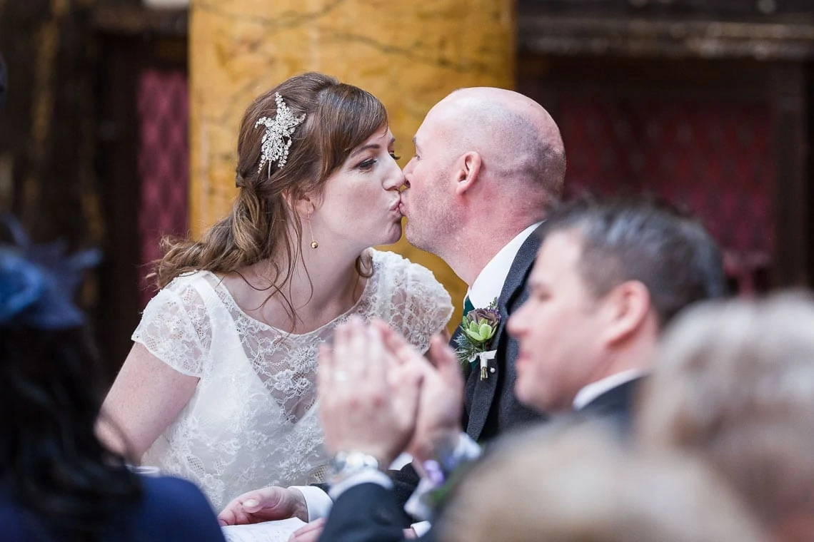 Bride and groom kissing during a wedding ceremony, with guests looking on and clapping in a decorated venue.