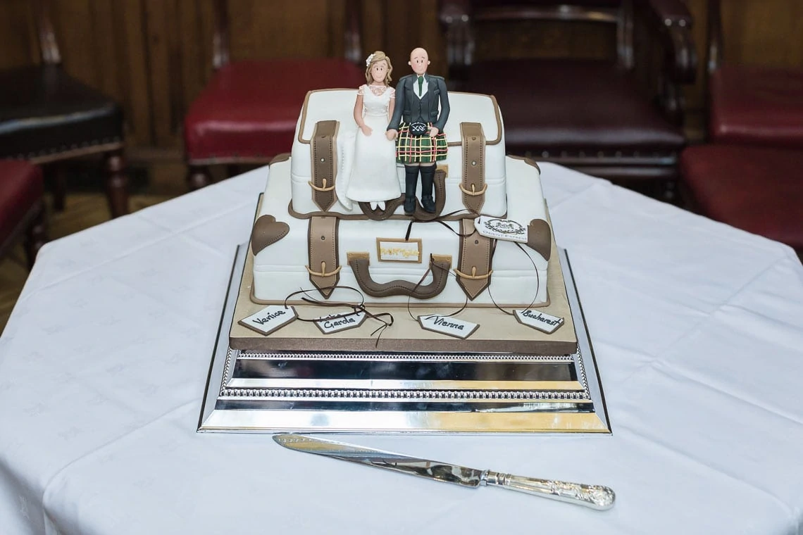 A wedding cake designed to resemble suitcases with a bride and groom figurine on top, displayed on a table with a cake knife beside it.