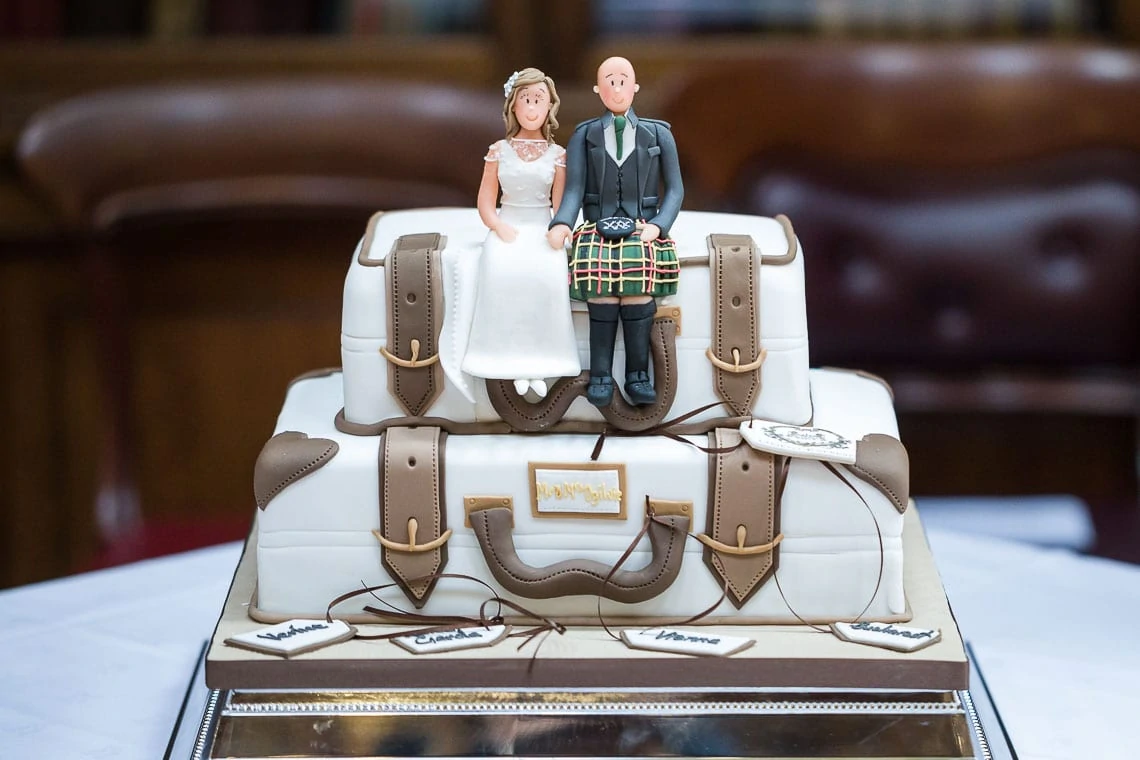 A wedding cake designed to look like stacked suitcases, topped with figurines of a bride and groom, the groom in a kilt.