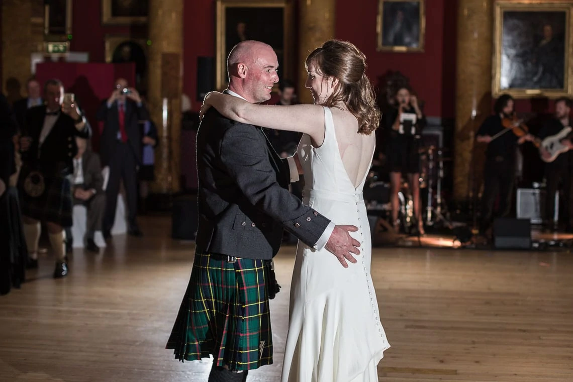 A couple dancing in a ballroom, the man in a kilt and the woman in a white dress, with a band playing in the background.