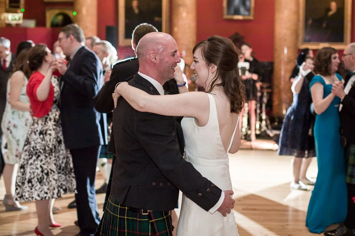 A man in a kilt and a woman in a white dress share a dance at a lively event with other guests dancing in the background.