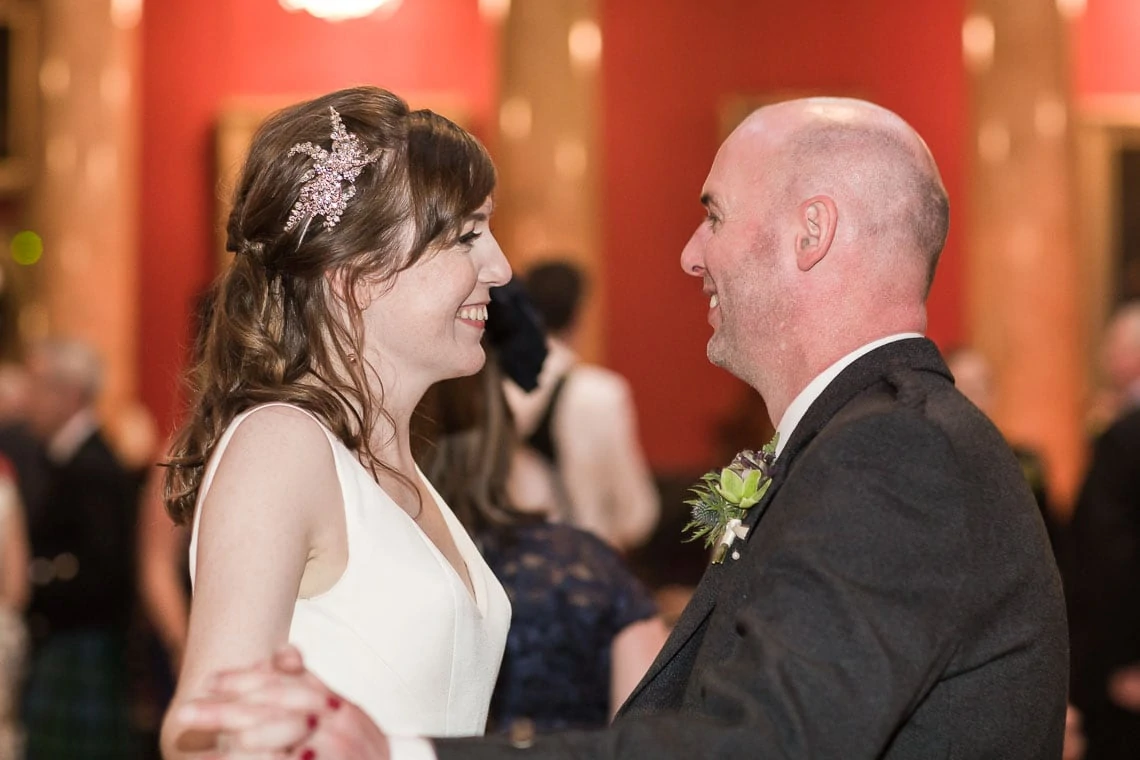 A bride and groom smiling at each other while dancing in a ballroom, surrounded by guests in formal attire.