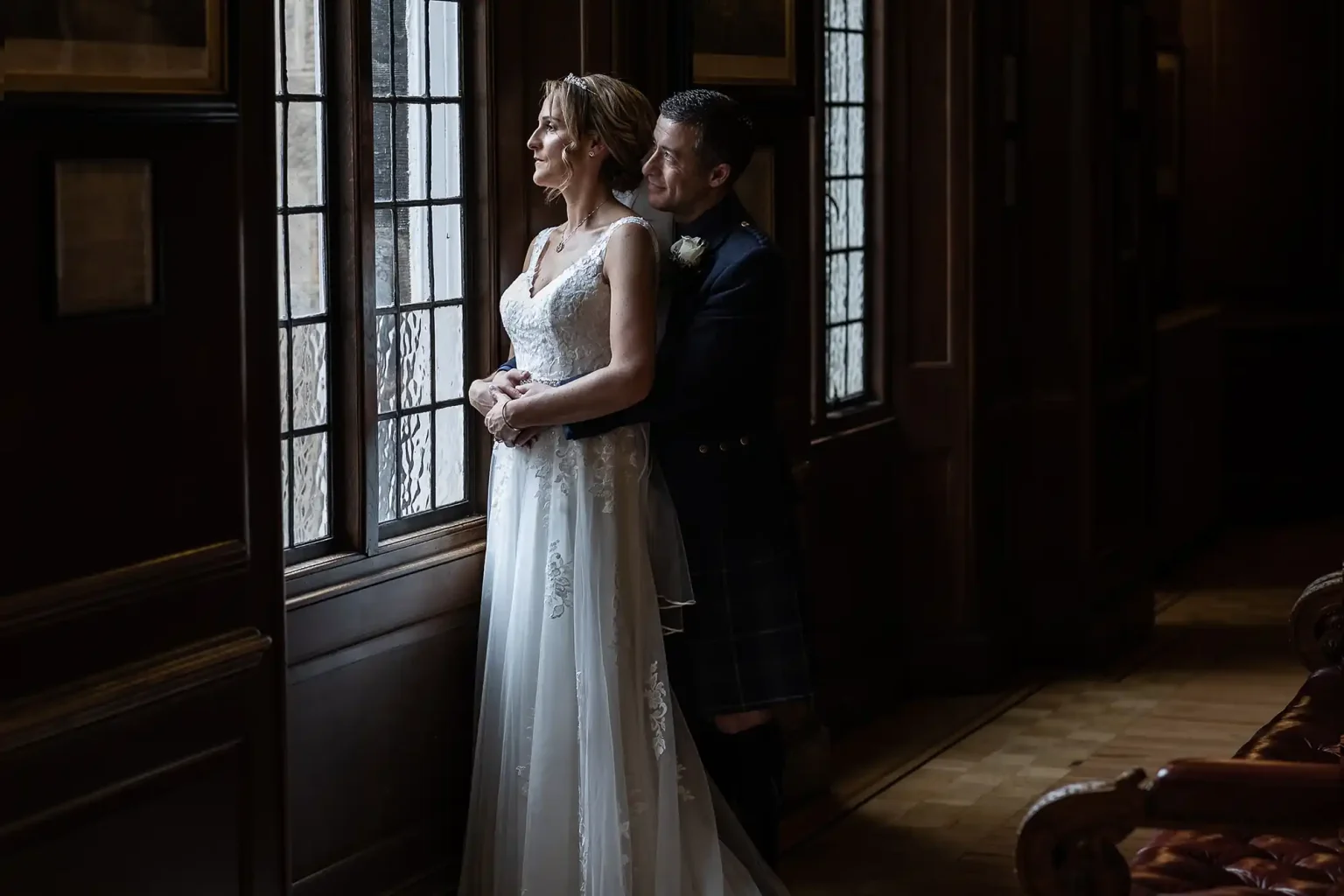 A bride in a white wedding dress and a groom in a dark suit stand gazing out of a window in a dimly lit room, with the groom holding the bride from behind.