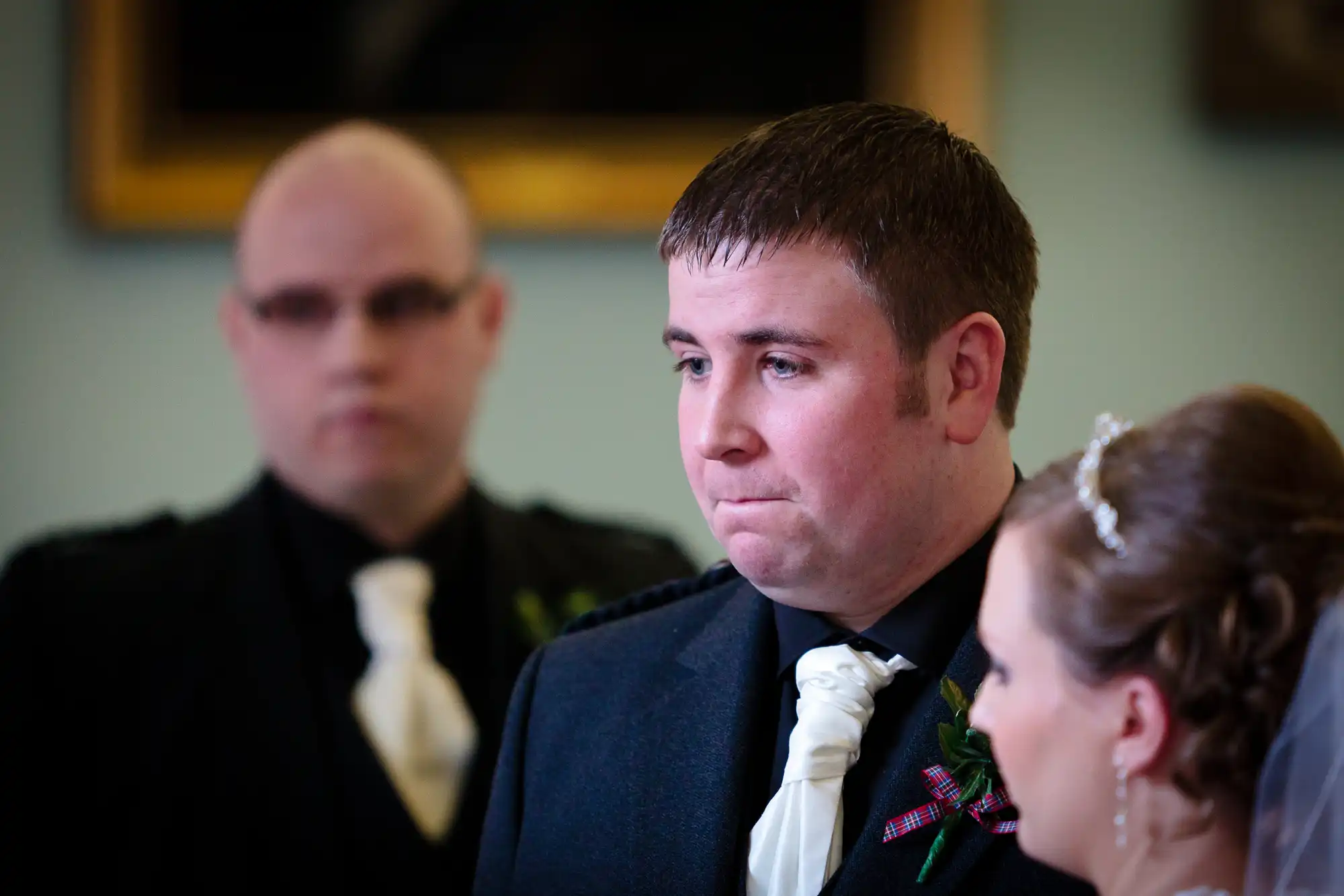 A groom in a dark suit with a tartan tie looks serious at a wedding ceremony, with a blurred figure in the background and a bride beside him.
