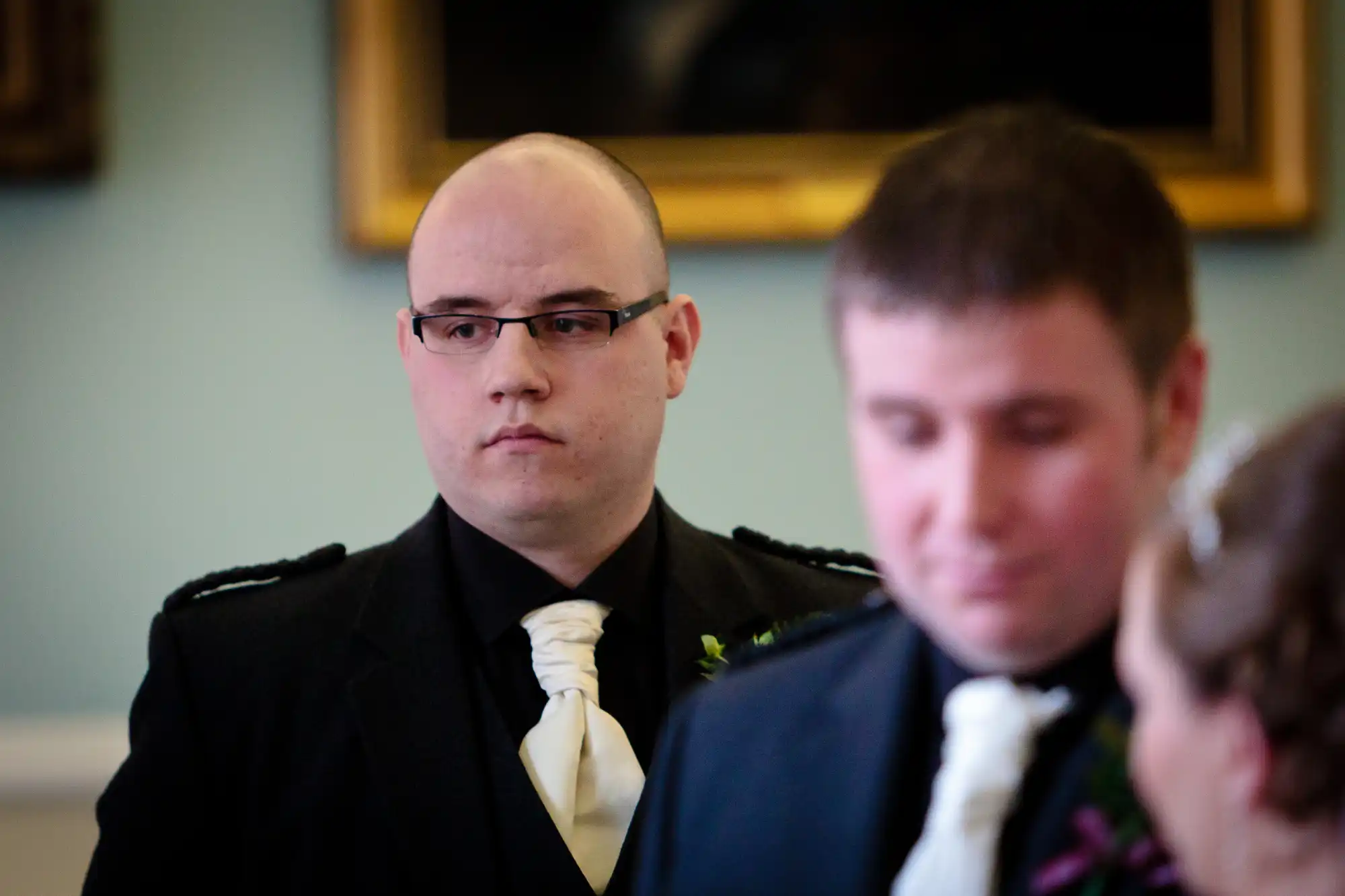 Bald groomsman in black suit and white tie, looking seriously at a wedding, with blurred bride and groom in the foreground.