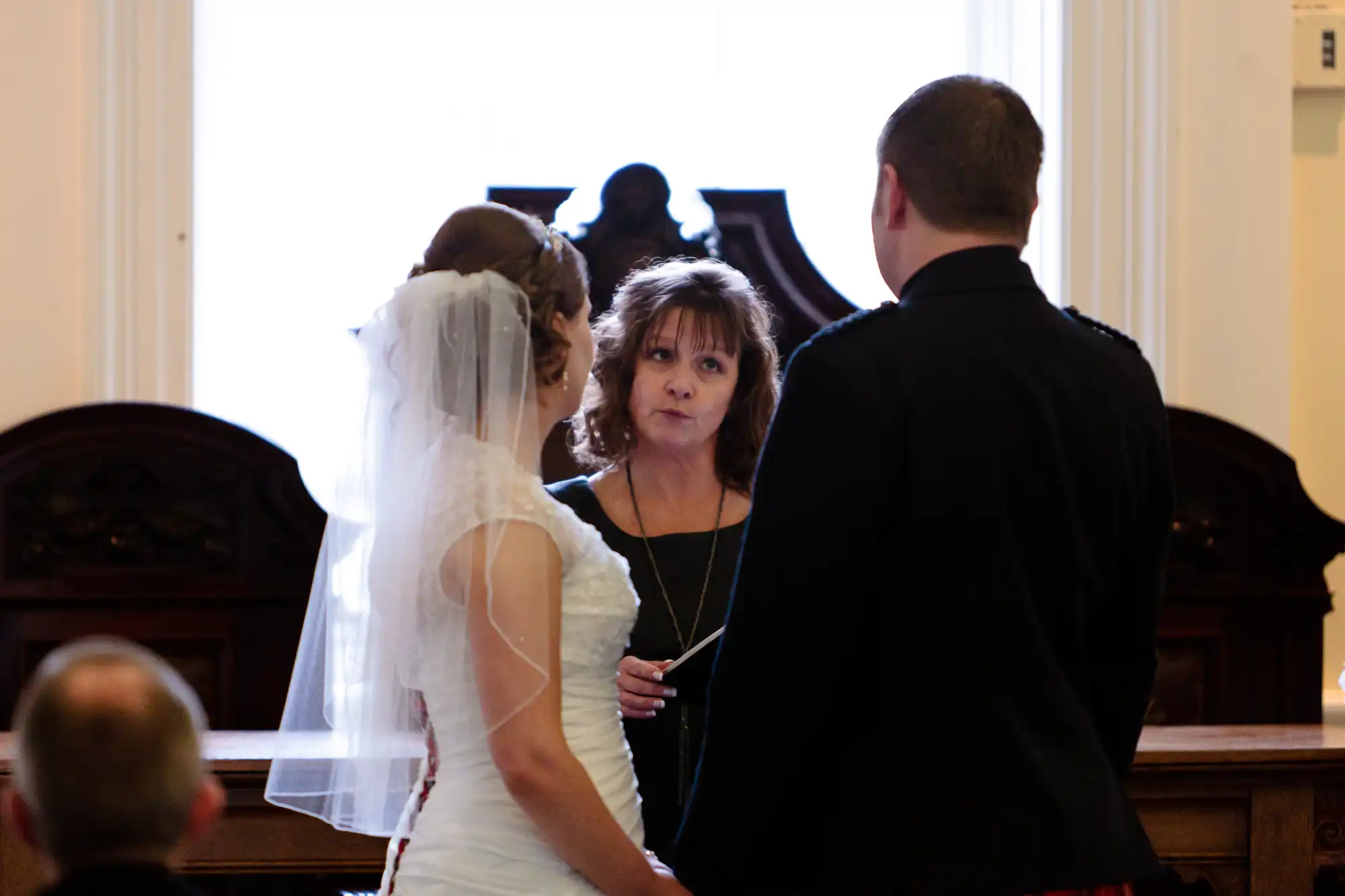 A wedding officiant looks at a bride and groom during a ceremony in an ornate room, viewed from behind the couple.