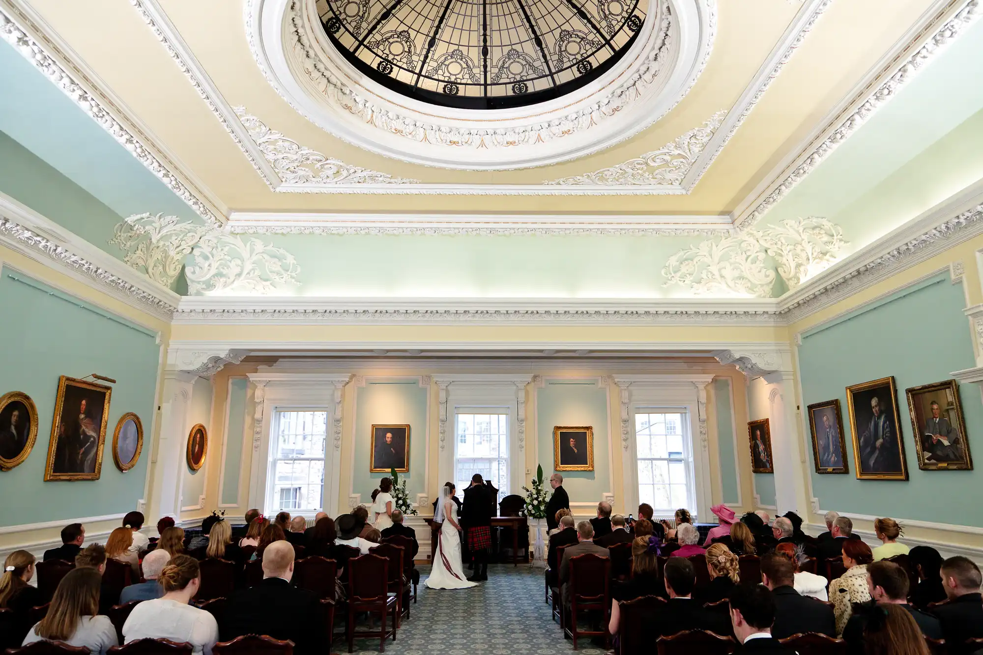 A wedding ceremony in an elegant room with a domed skylight, pastel walls, decorative moldings, and portraits, as guests seated in rows watch a couple at the altar.