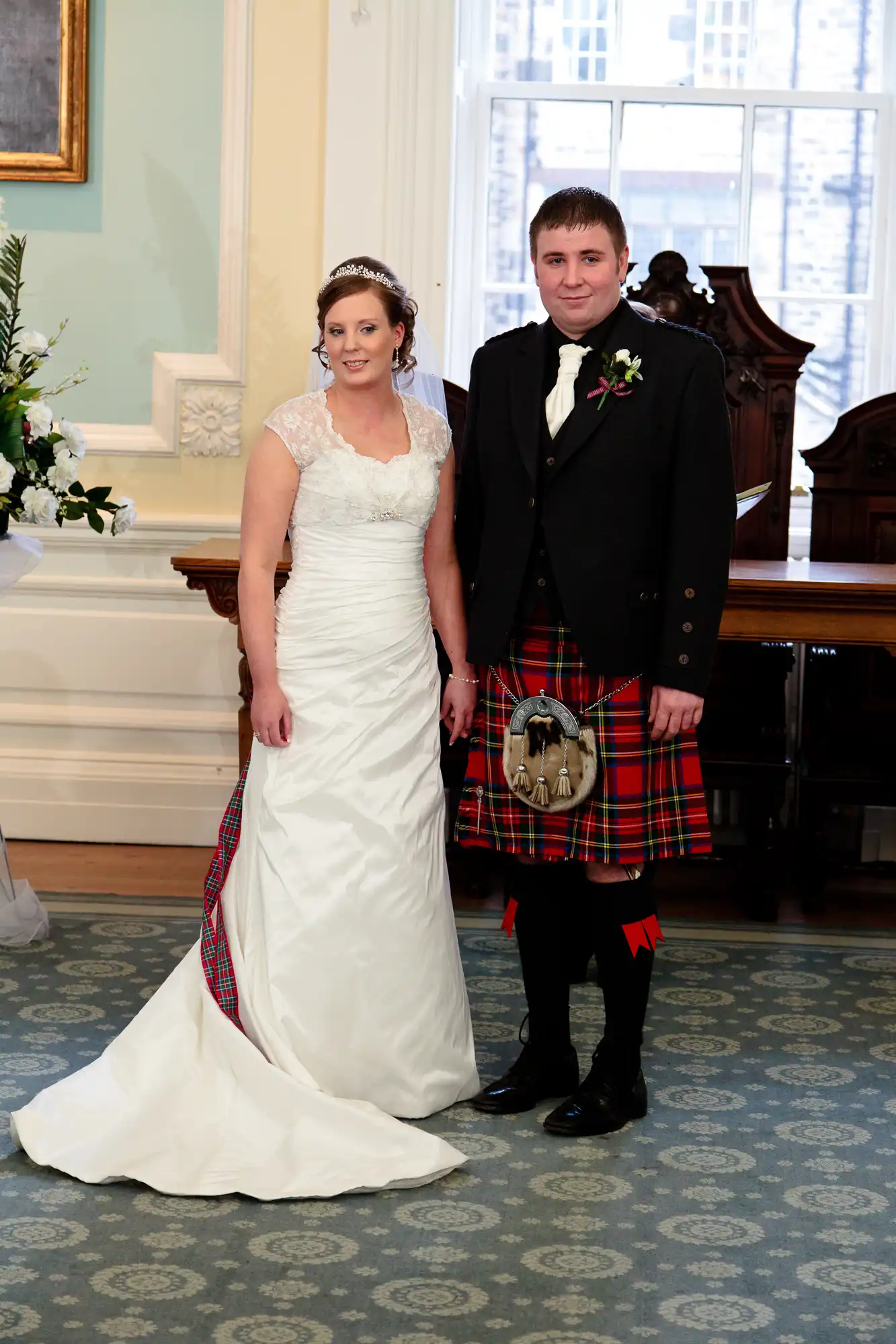 A bride in a white gown and a groom in a tartan kilt and sporran standing in a room with elegant decor.