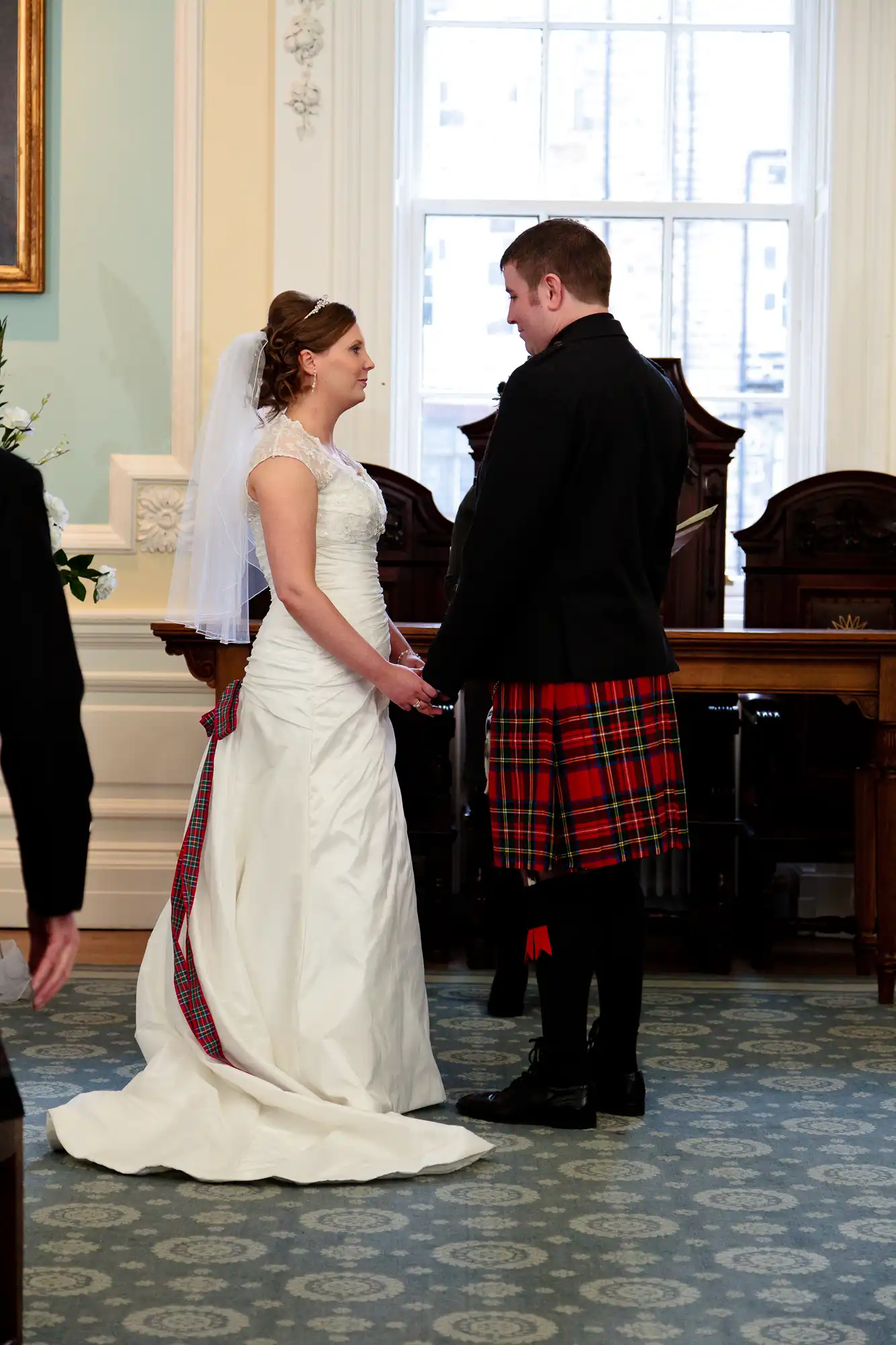 A bride in a white gown and a groom in a tartan kilt hold hands at their wedding ceremony inside a room with traditional decor.
