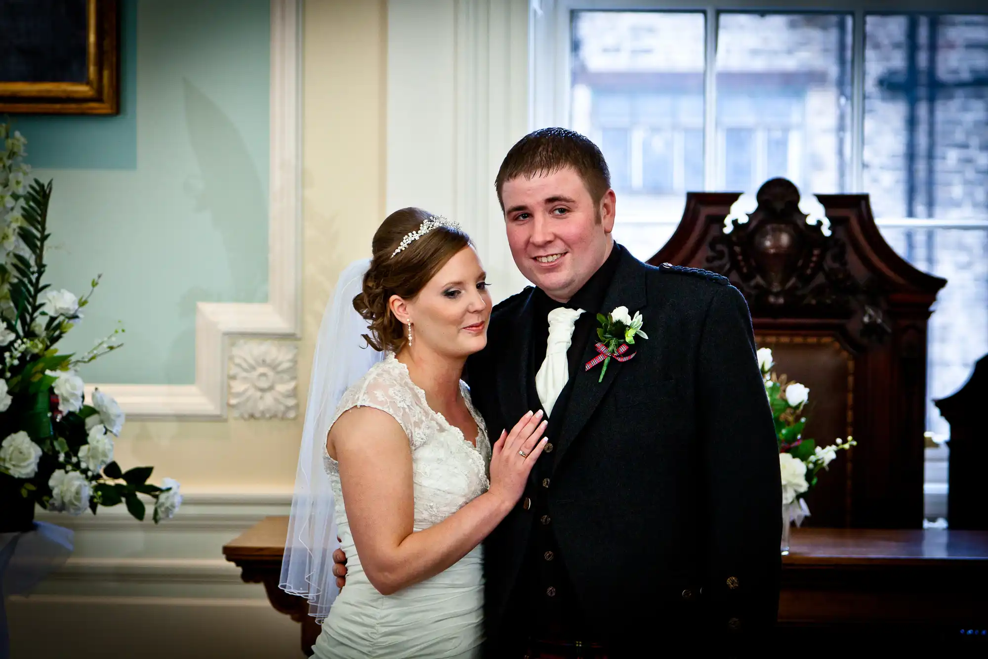 A bride and groom smiling in a classic indoor setting, the bride in a white dress and the groom in a suit and kilt.