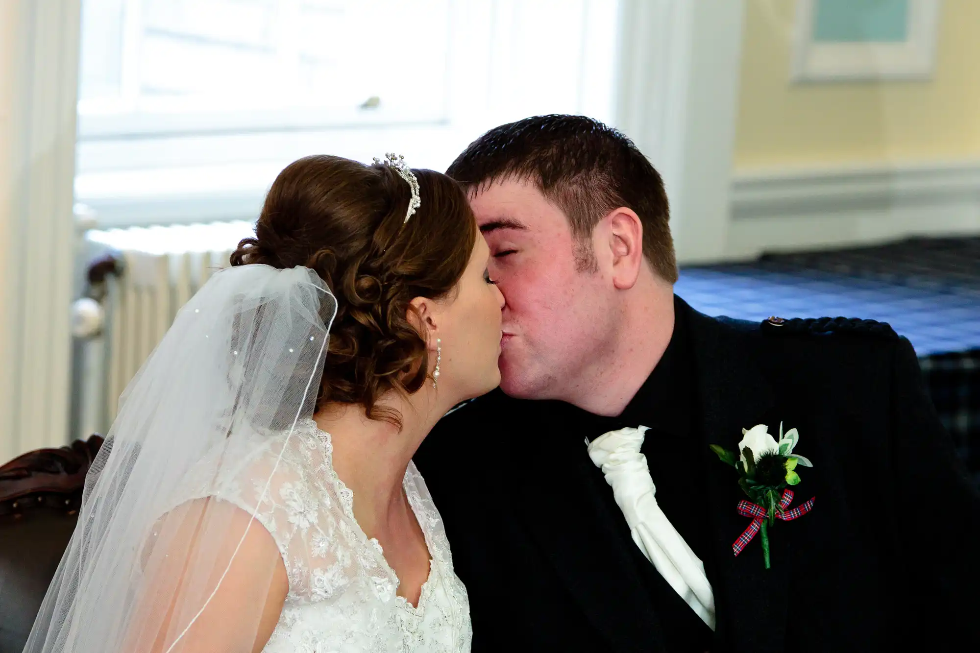 A bride and groom kissing, dressed in traditional wedding attire, indoors with natural light coming through windows in the background.