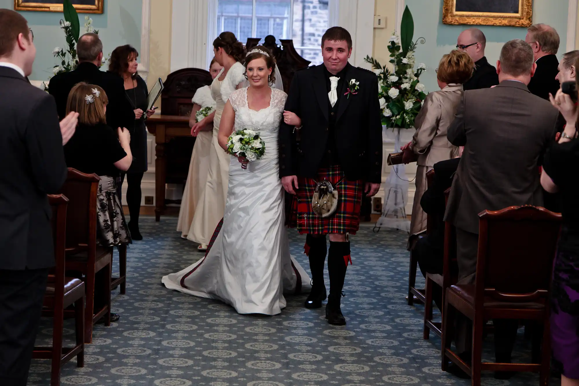 Bride in a white dress and groom in a kilt walking down the aisle, smiling, with guests applauding in a decorated room.