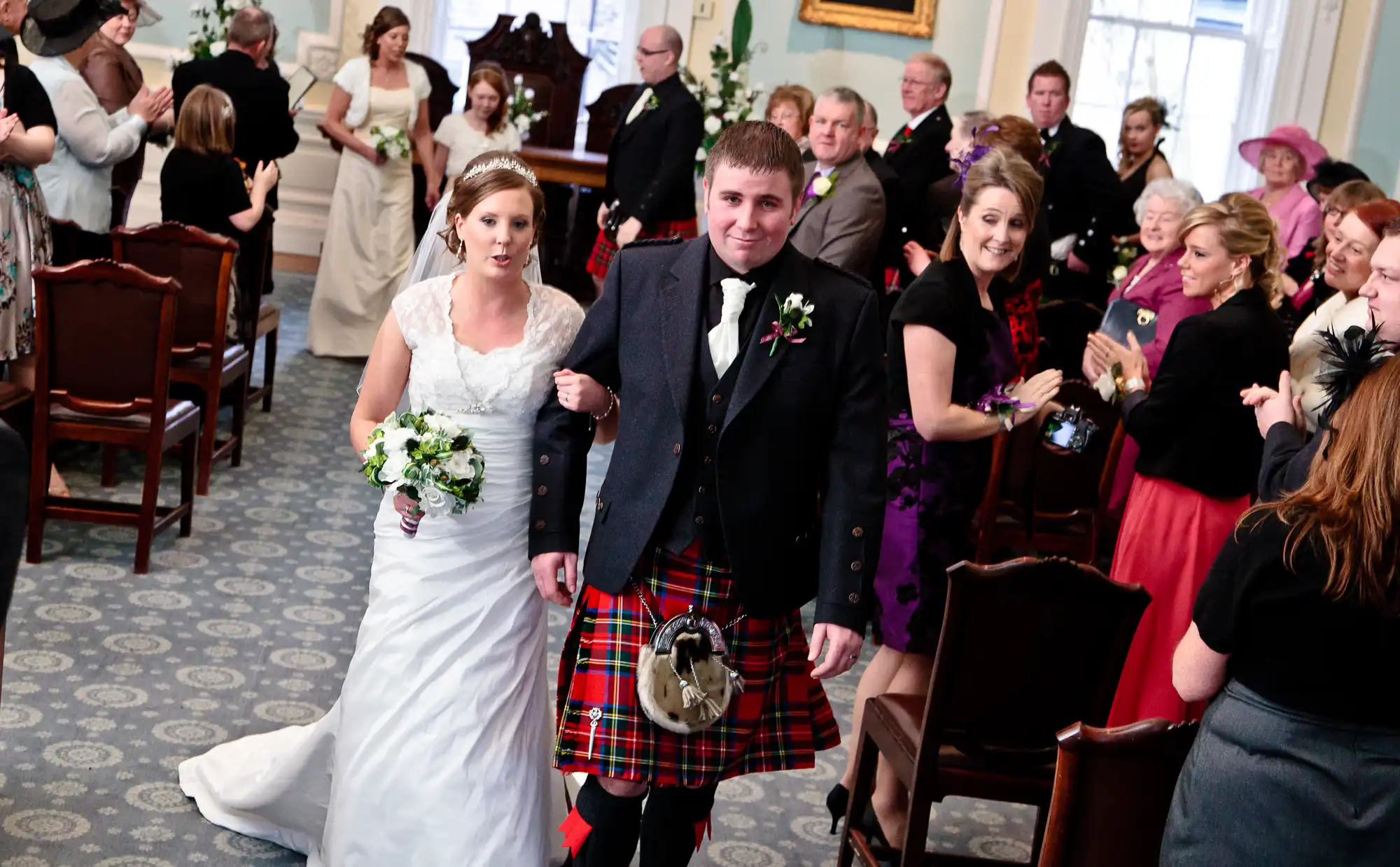A bride in a white dress and a groom in a kilt walk down the aisle, surrounded by applauding guests in a decorated room.