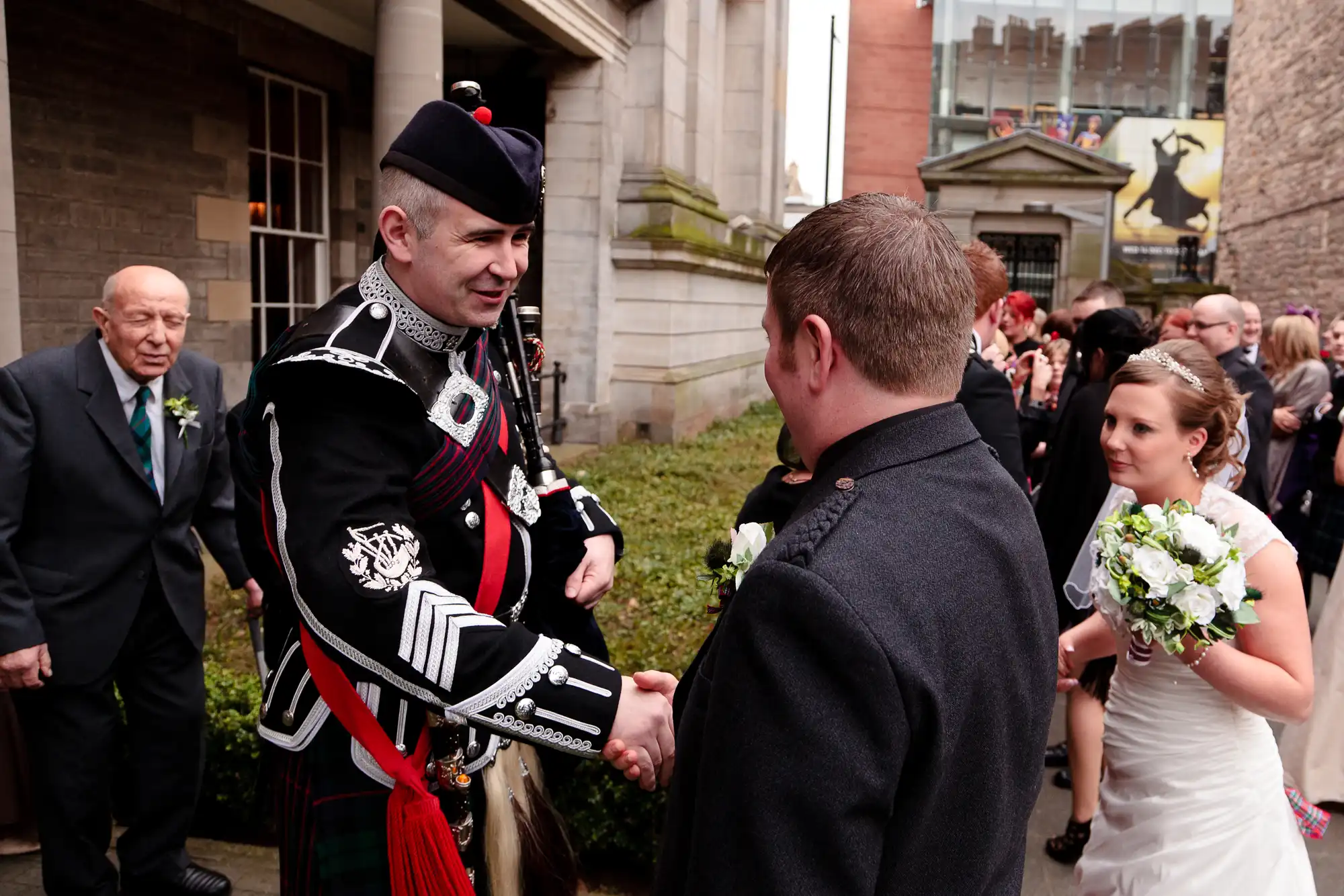 A bride looks on as a man in a scottish kilt shakes hands with another man at a wedding ceremony outside a building.