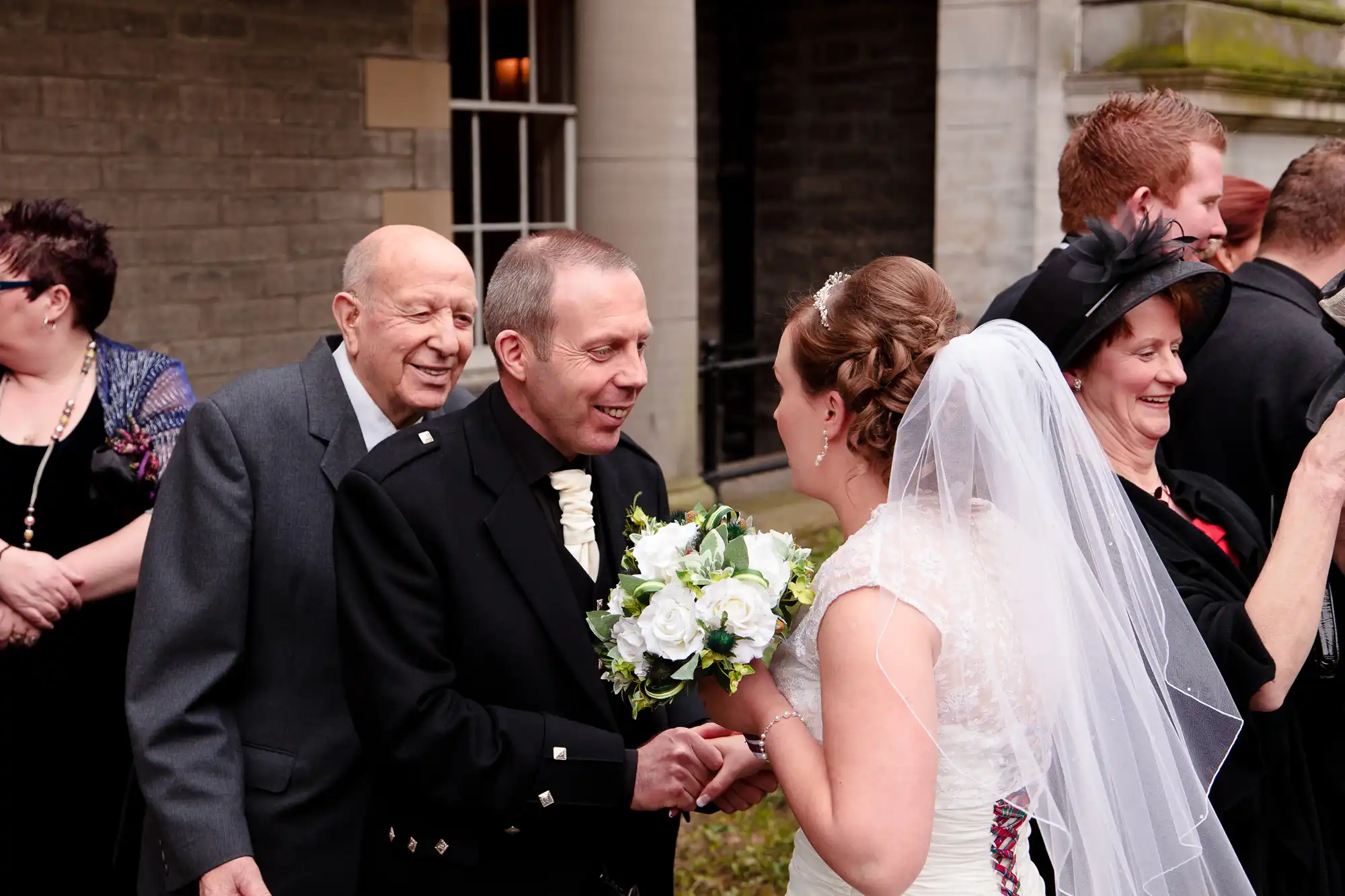 A bride and groom shake hands with an elderly man at a wedding, surrounded by smiling guests.