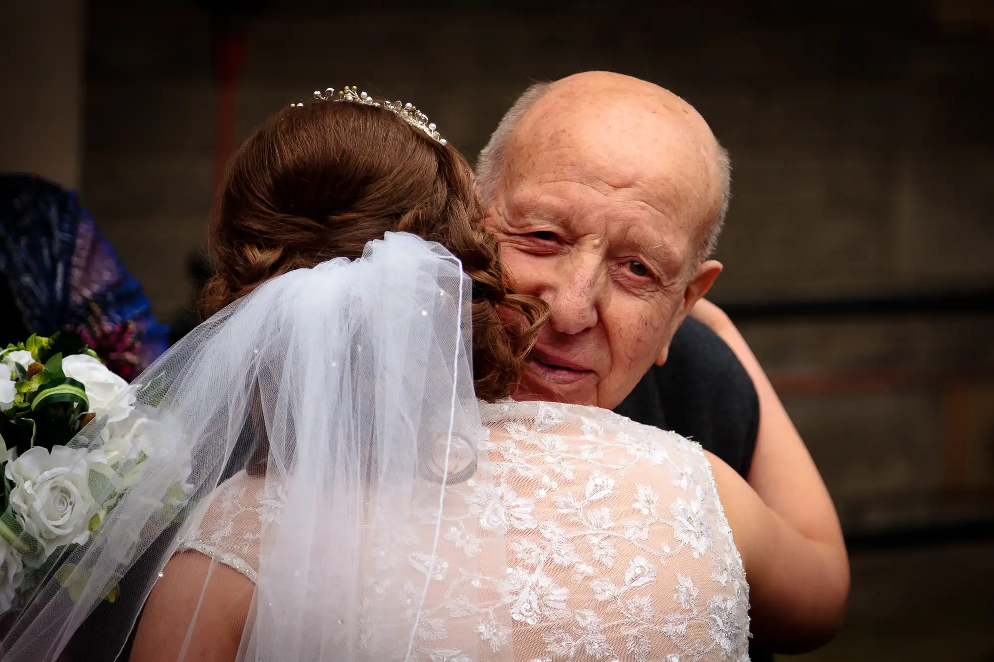 Elderly man emotionally embracing a bride in a lace wedding dress and veil, holding a bouquet, in an outdoor setting.