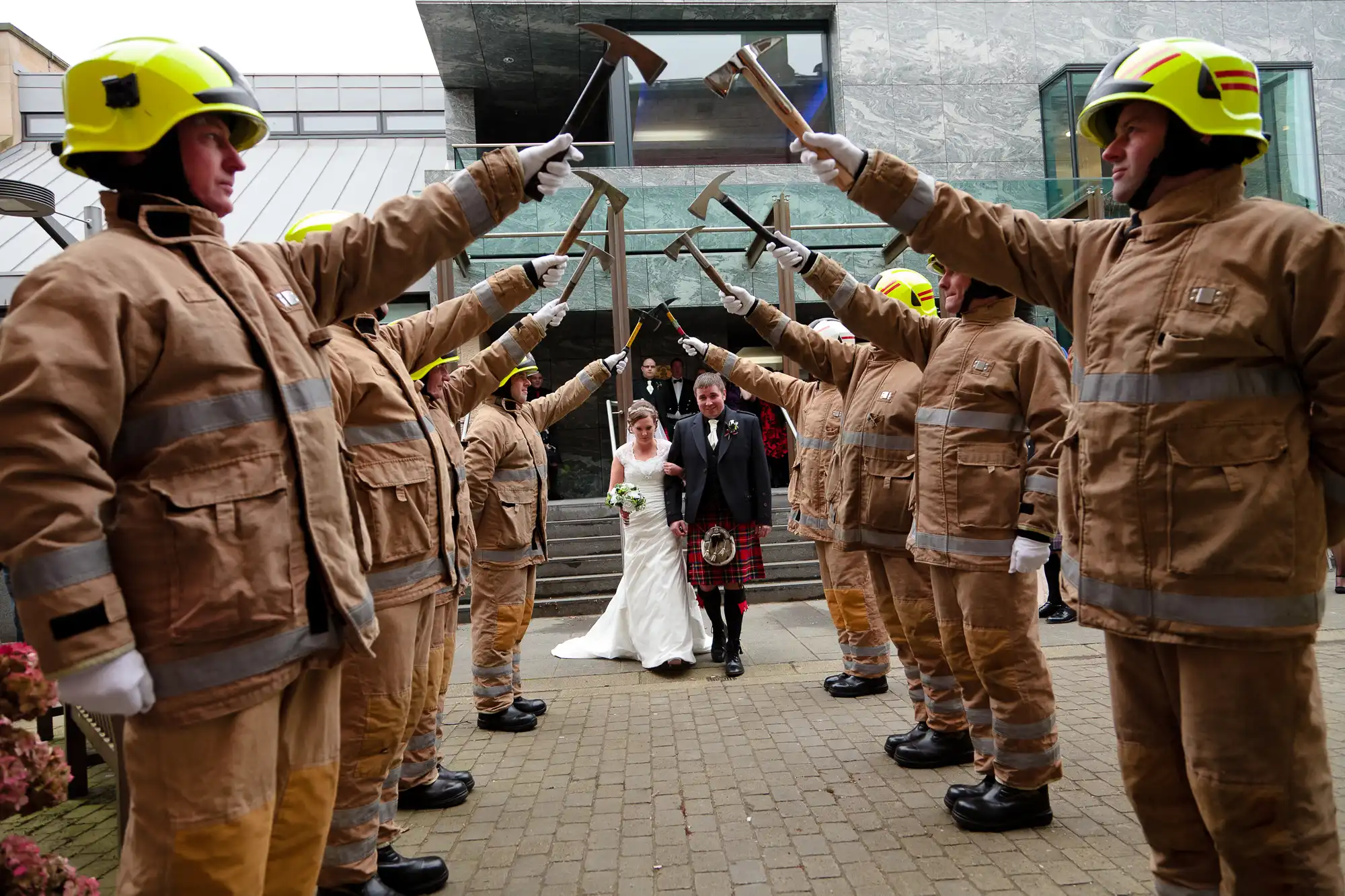 A bride and groom walk under an arch of axes held by firefighters in uniform, celebrating their wedding outside a building.