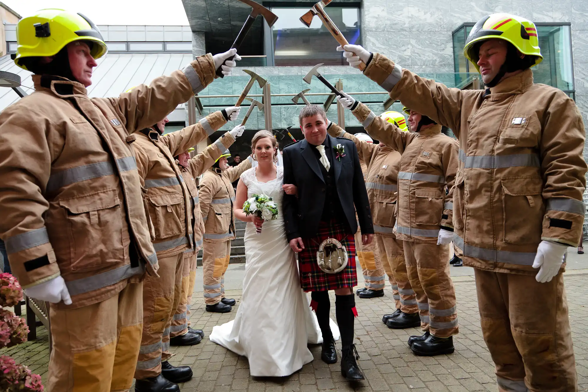 A bride and groom walk through an archway made by firefighters holding axes, outside a building on a foggy day.