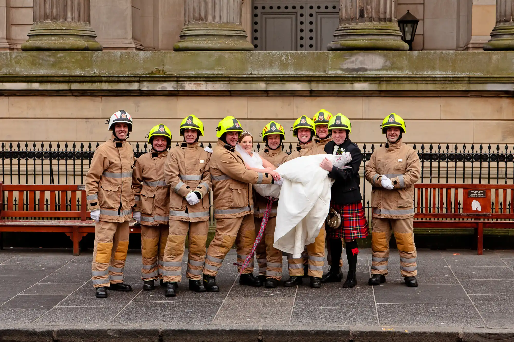 A group of eight firefighters in full gear, including one in a kilt, posing with a bride in front of a bench on a city street.